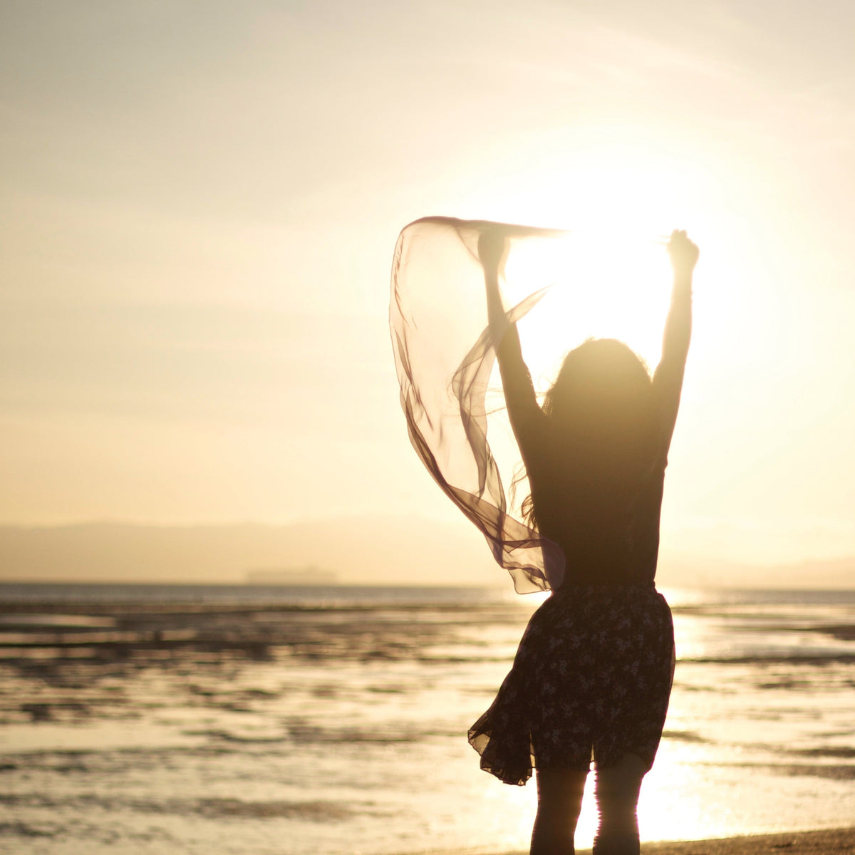 Silhouette of a woman on a beach at sunset holding a sheer scarf above her head