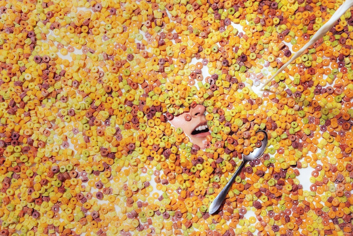 A smiling person's face peeking out from a pile of colorful ring cereal, with a spoon and milk being poured