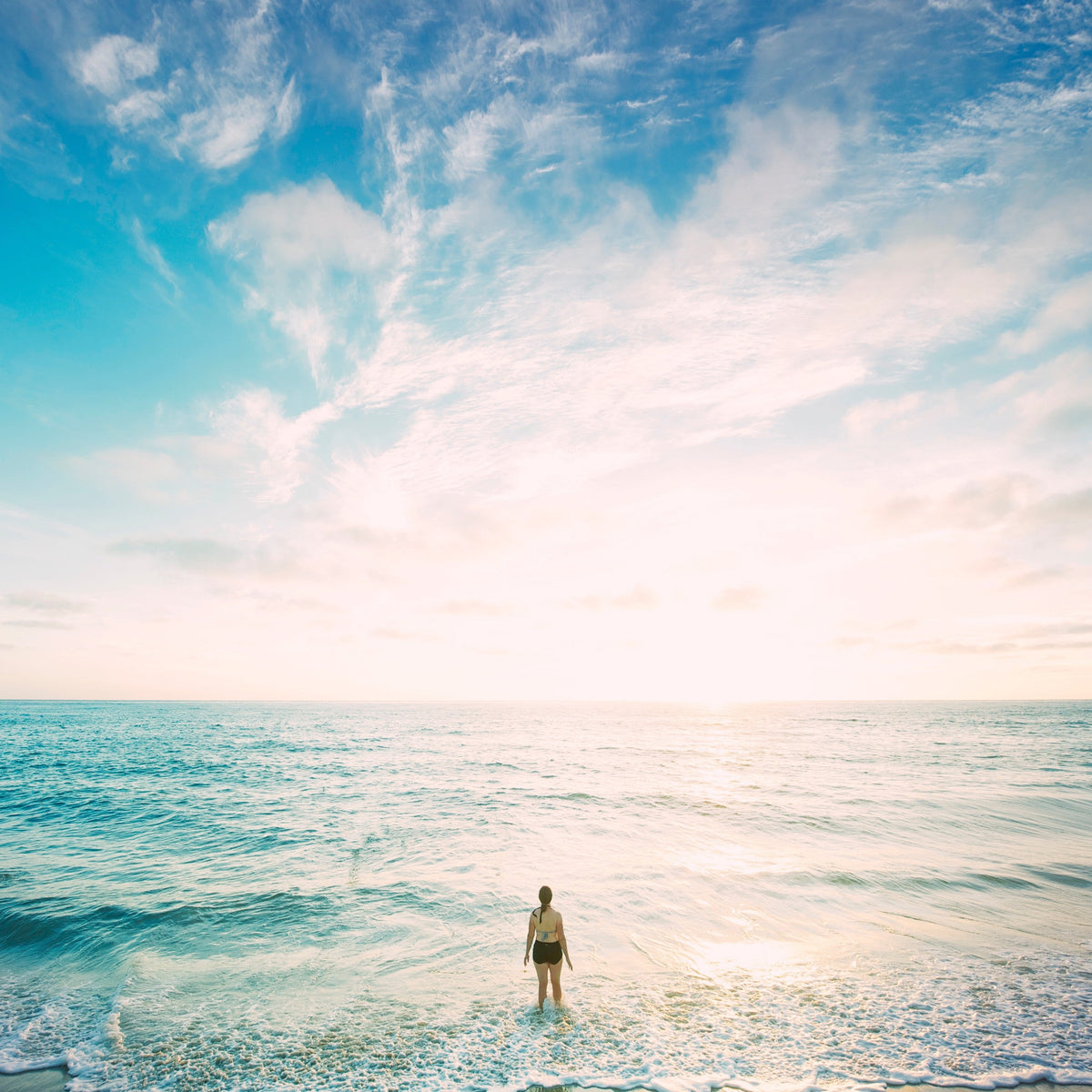 A person standing in shallow ocean water, looking out at the horizon under a bright sky