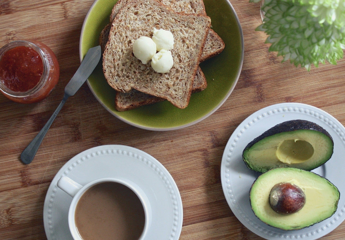 Overhead view of whole-grain toast with butter and jam, a cup of coffee and a halved avocado