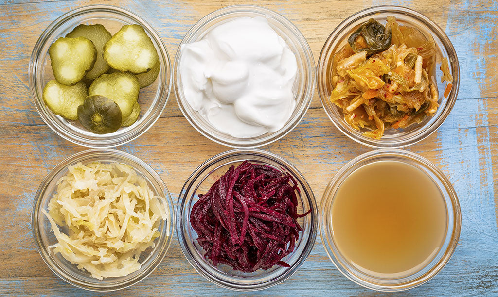 Six glass bowls of fermented foods, including pickles, yogurt, kimchi, sauerkraut, pickled beets and a fermented drink
