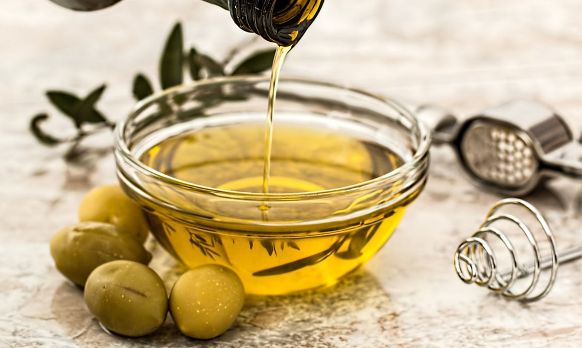 Olive oil being poured into a glass bowl beside green olives