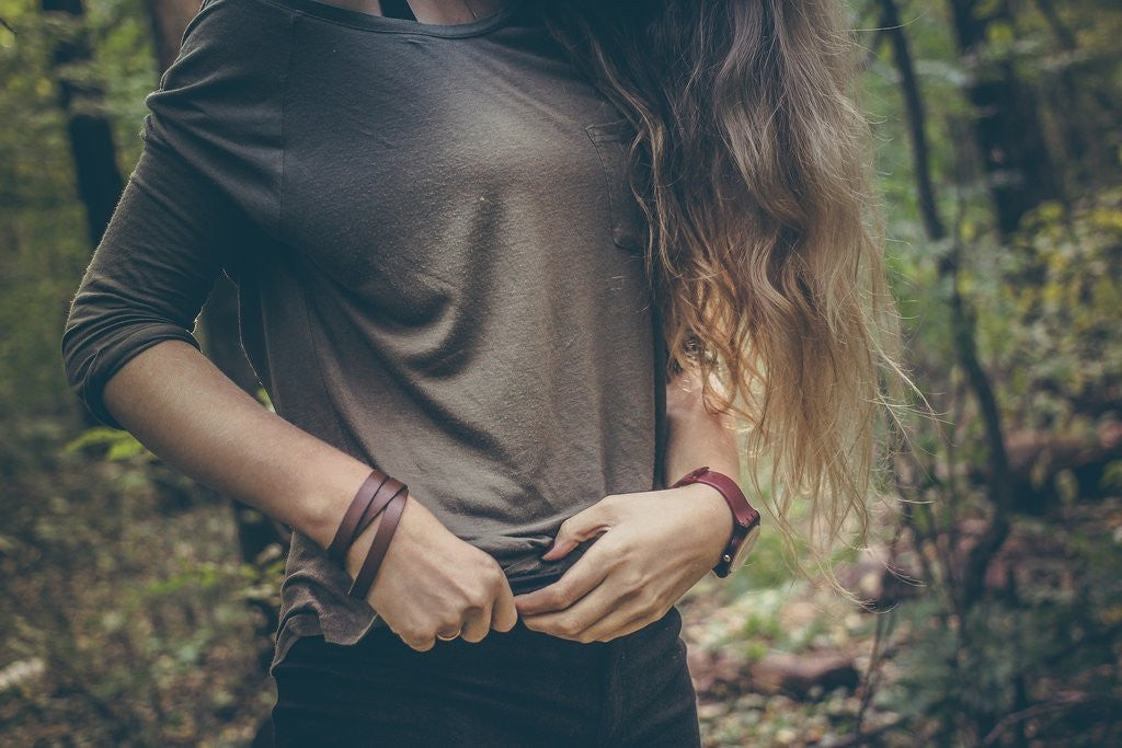 Woman in a brown top standing in a forest, holding the hem of her shirt near her stomach