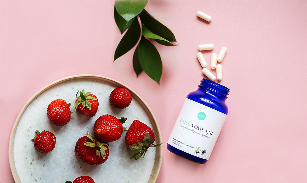 Bottle of Trust Your Gut probiotic capsules with loose capsules, strawberries and green leaves on a pink background