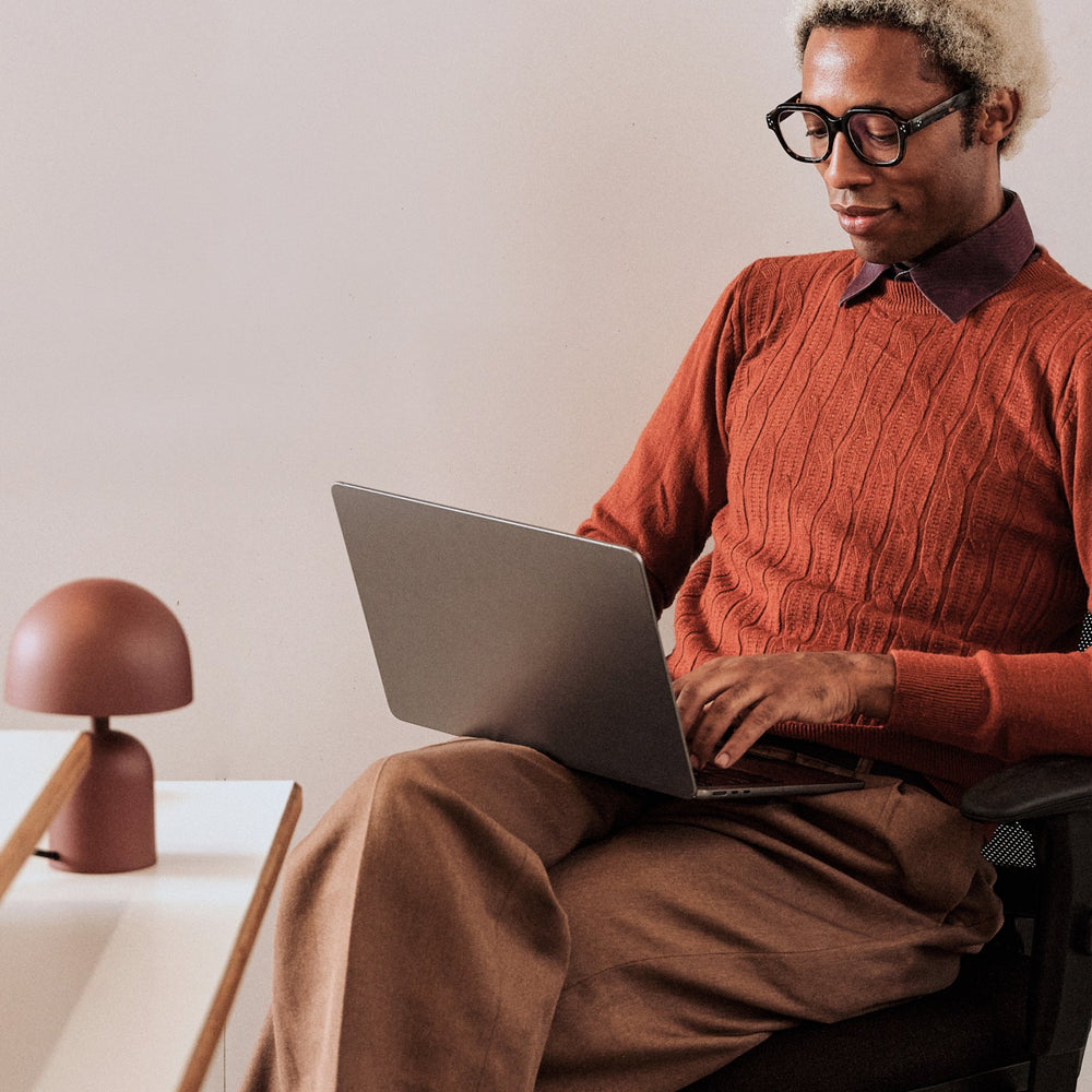A man in an orange sweater working on a laptop at his desk.