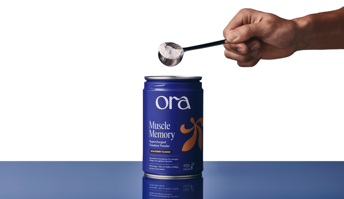 A hand holding a scoop of white powder over a blue can of Ora Muscle Memory creatine.