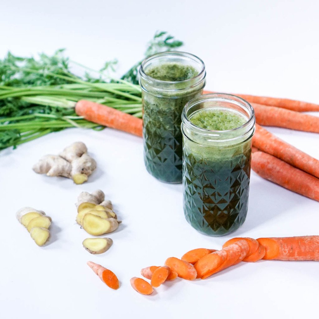 Two jars of green juice with carrots and ginger on a white surface.